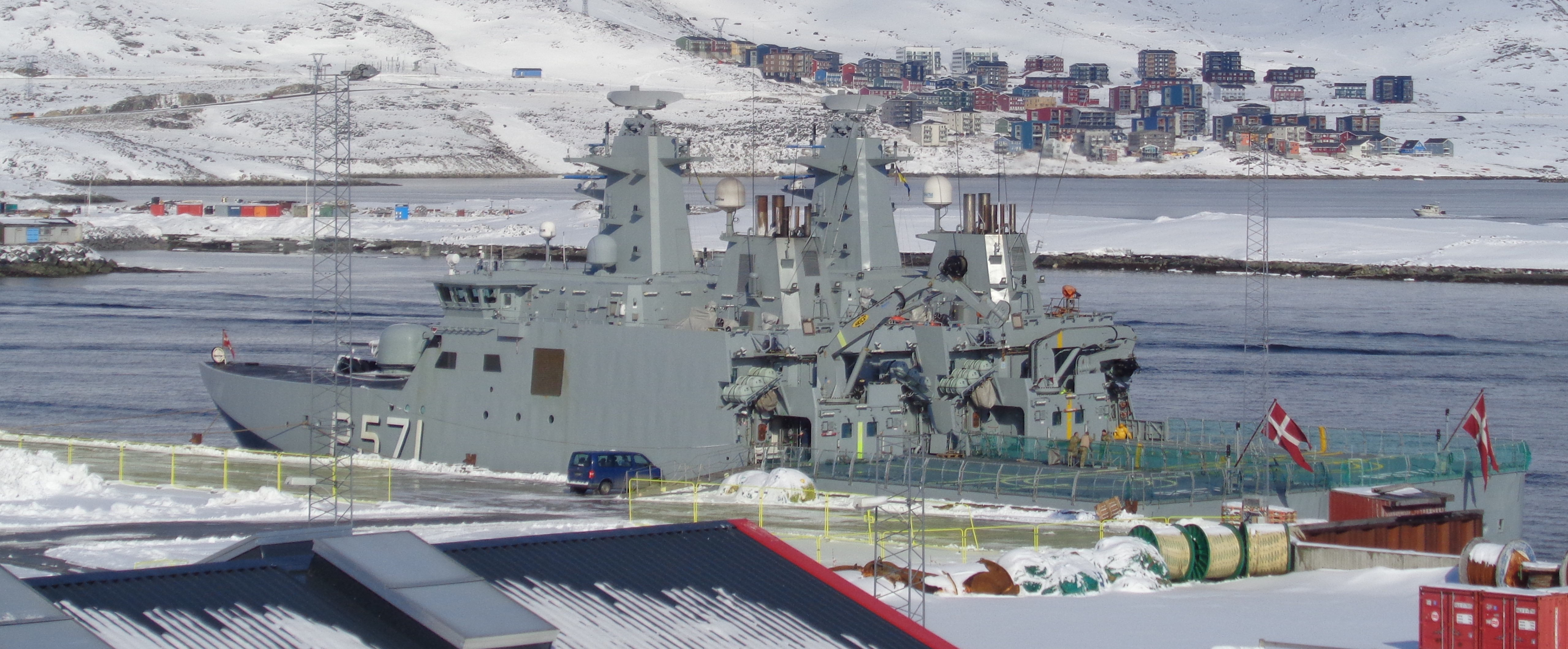 Gray Danish Royal Navy vessel marked "5571" docked in port at Nuuk with a snowy waterfront and hillside buildings behind it.