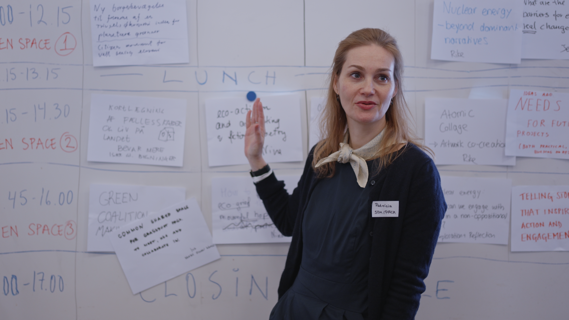 Woman with a name tag reading "Patricia SDN/IPCC" speaking and gesturing beside a wall covered with handwritten workshop notes, including "LUNCH," "Atomic Collage," and "Nuclear energy - beyond dominant narratives."