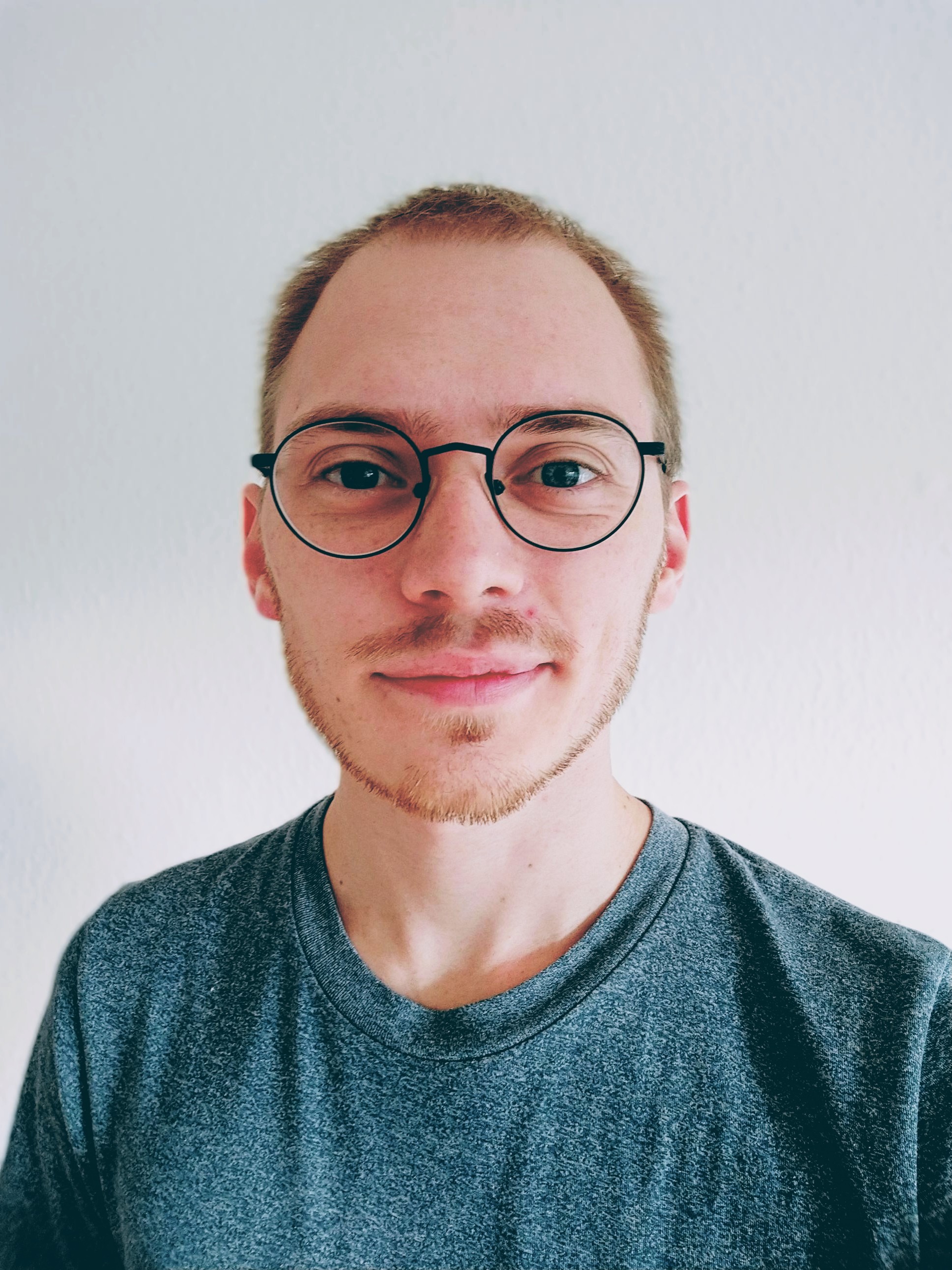Tæt portræt af Kasper Friis Bavnbaek med runde briller og grå T-shirt foran en lys baggrund.
