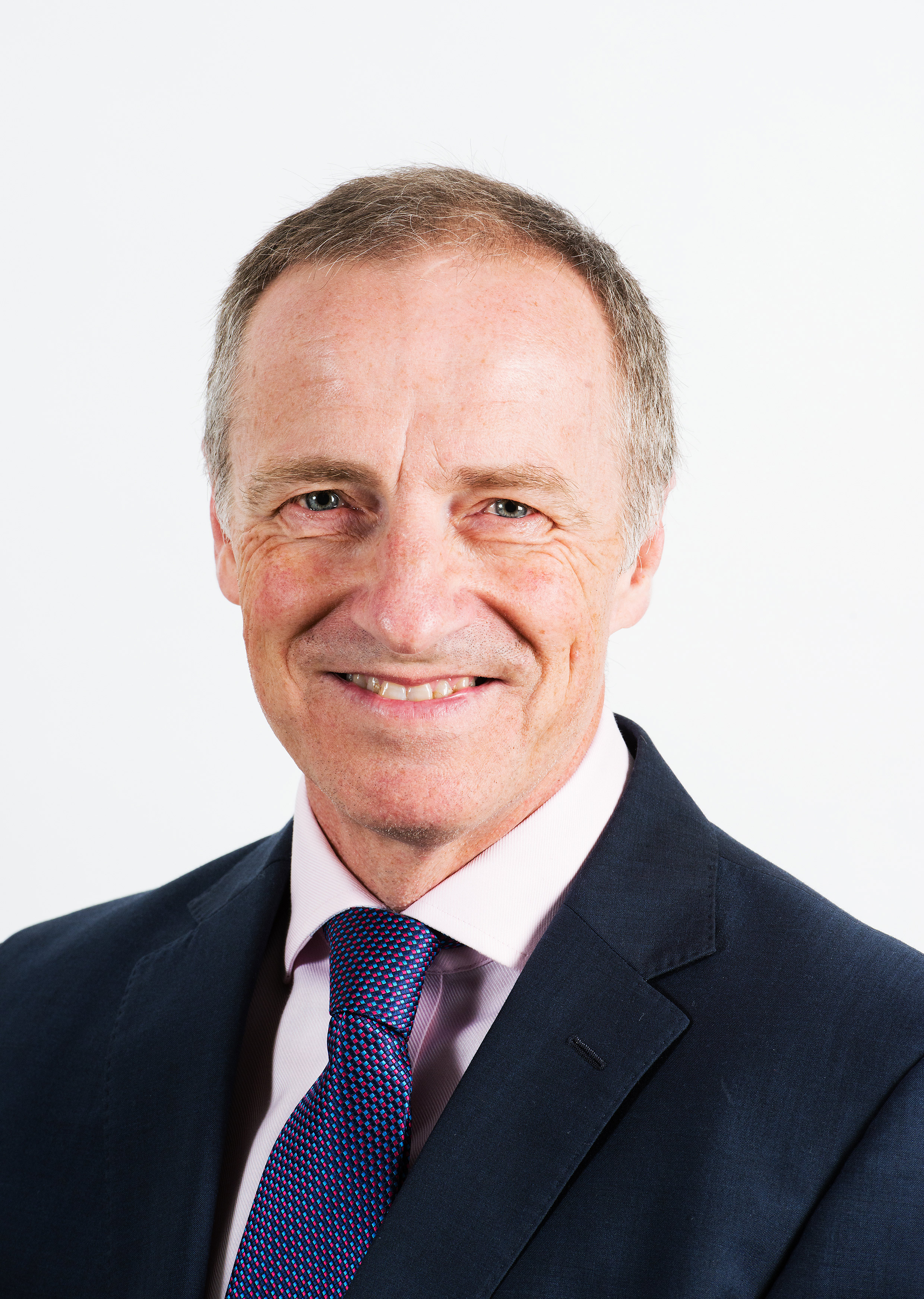 Professional portrait of Alastair Benbow in a dark suit and patterned tie, smiling against a plain light background.