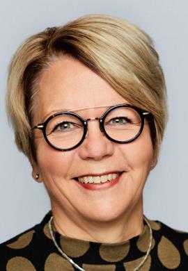 Portræt af Benedicte Vibjerg Wilson med kort blondt hår, runde briller og et smil mod en grå baggrund.