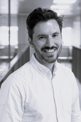 Black-and-white portrait of a smiling bearded man in a light button-down shirt standing in an office corridor.