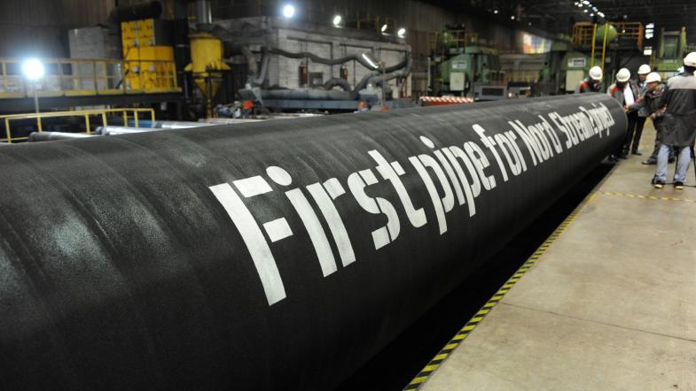 Stort sort industrirør i en fabrikshal med teksten "First pipe for Nord Stream 2 ha" og arbejdere i baggrunden.