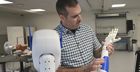 Robotforsker, Oskar Palinko, står med en humanoid robot og undersøger dens ene hånd.