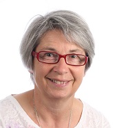 Sue Charlesworth smiling in a head-and-shoulders portrait, wearing red glasses against a light gray background.