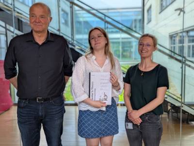 Three people posing indoors, with the center person holding a printed document.