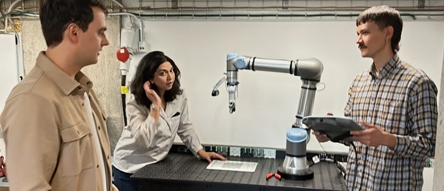 Three people observing and operating a robotic arm at a lab worktable.
