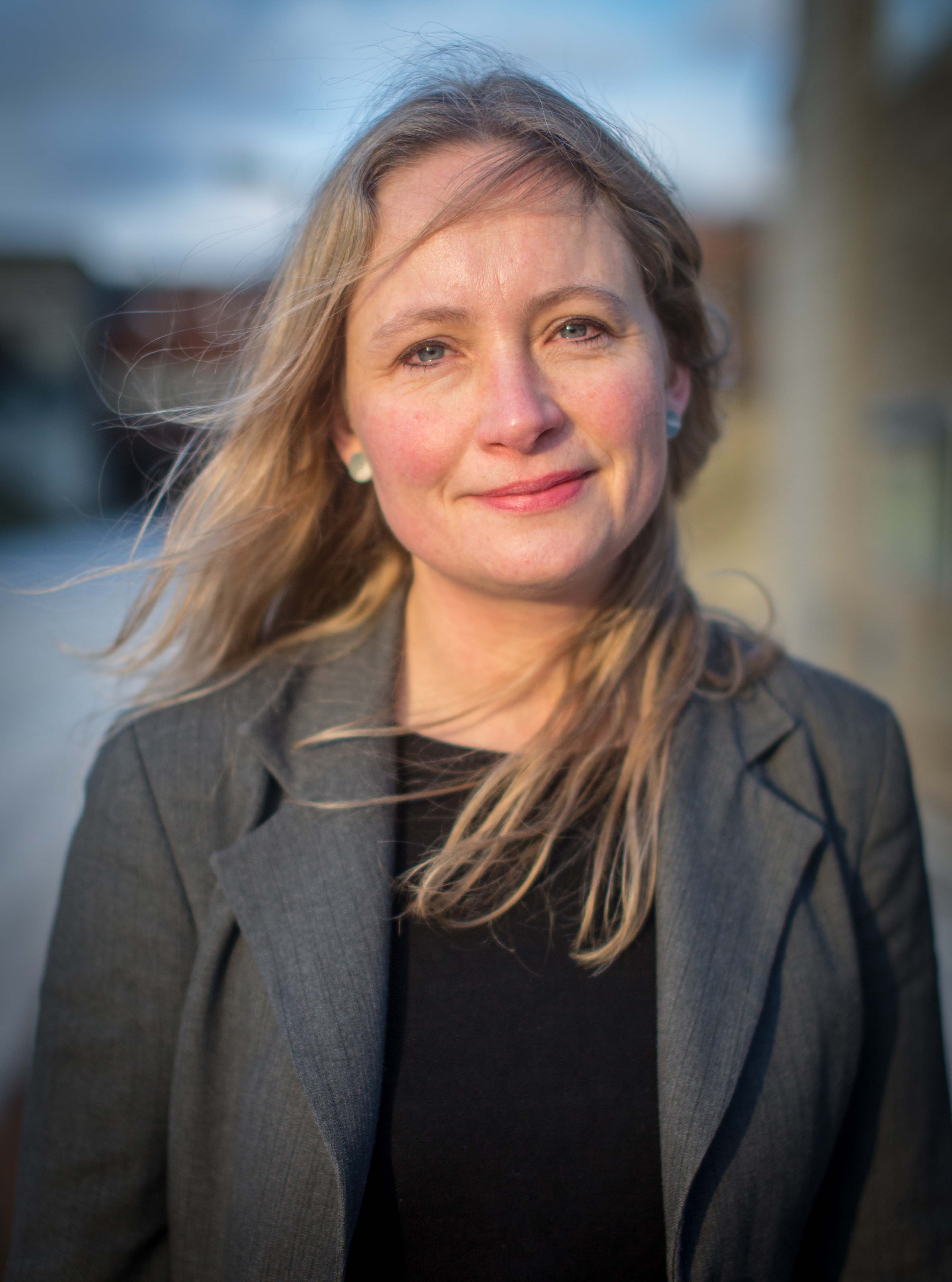 Kvinde med lyst hår i grå blazer fotograferet udendørs med sløret baggrund.