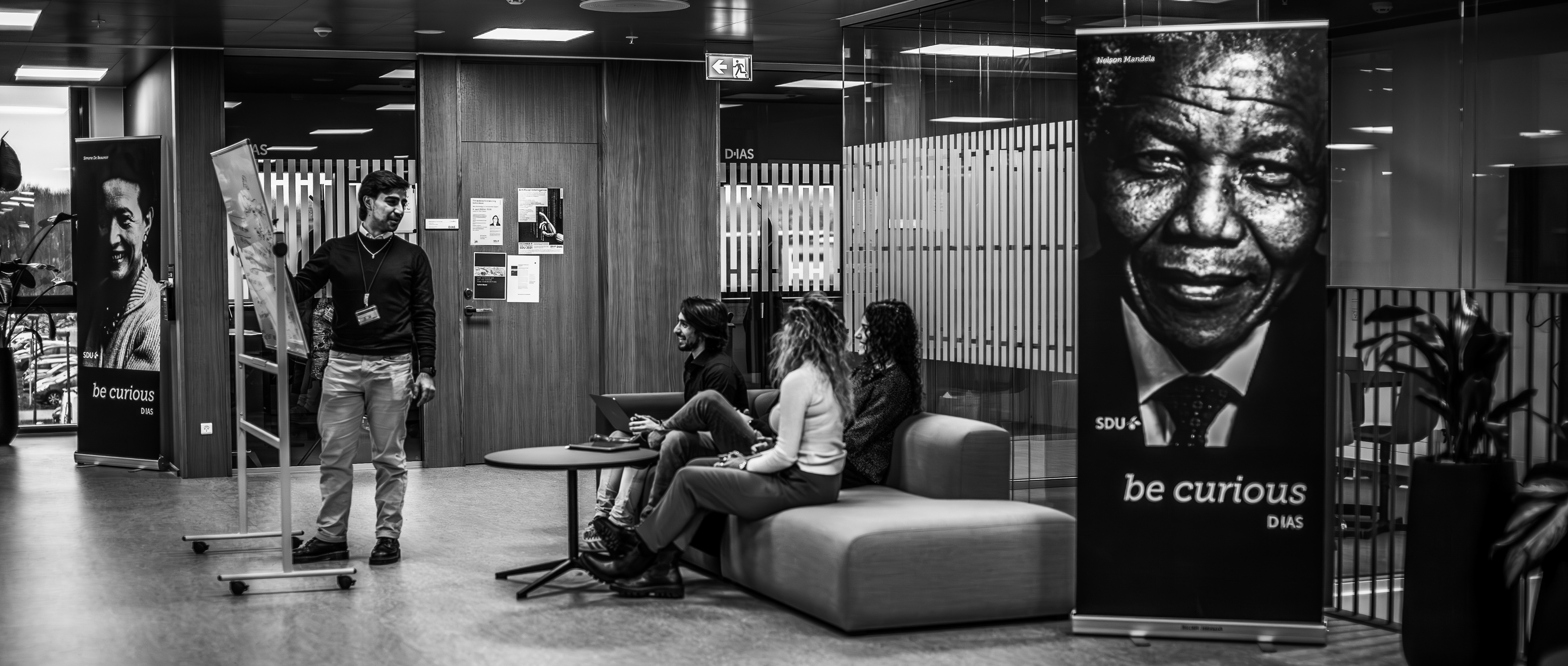 Fire unge mennesker i DIAS-bygningen. En står ved er whiteboard og fortæller, mens tre sidder i en sofa og lytter.