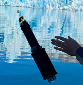 A hand catches a cylindrical water-sampling device into calm water near the blue-white face of a glacier, with the ice reflected on the surface.