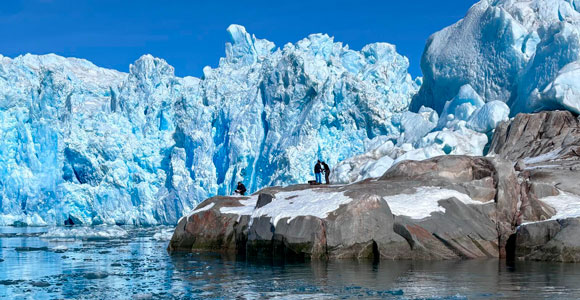 Several researchers stand on rocky ground at the edge of a glacier, working with a drone, with ice formations and cold water in the background.
