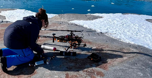 A woman works by a drone, which is placed on a cliff by the icy waters in the Arctic