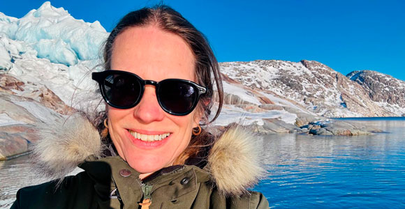 A person takes a selfie in an Arctic landscape, wearing sunglasses and a warm jacket, with ice, mountains, and calm water in the background beneath a clear blue sky.