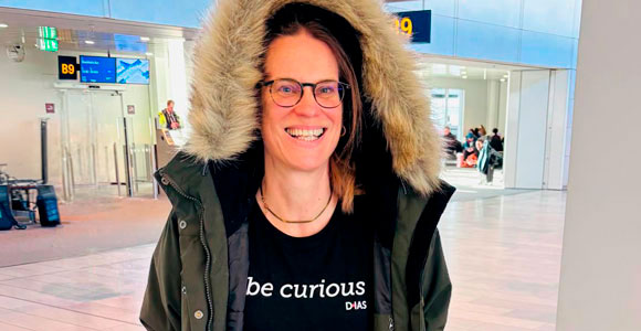 A person is standing in an airport wearing a winter jacket with a hood and glasses. The text on the sweater reads “be curious DIAS”.