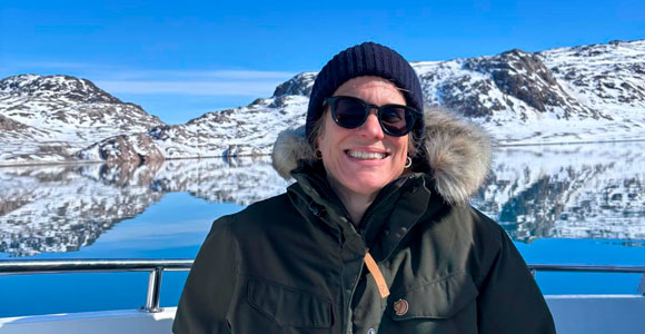 A researcher stands on a boat in an Arctic landscape, wearing warm outerwear and sunglasses, with snow-covered mountains and glassy water in the background.