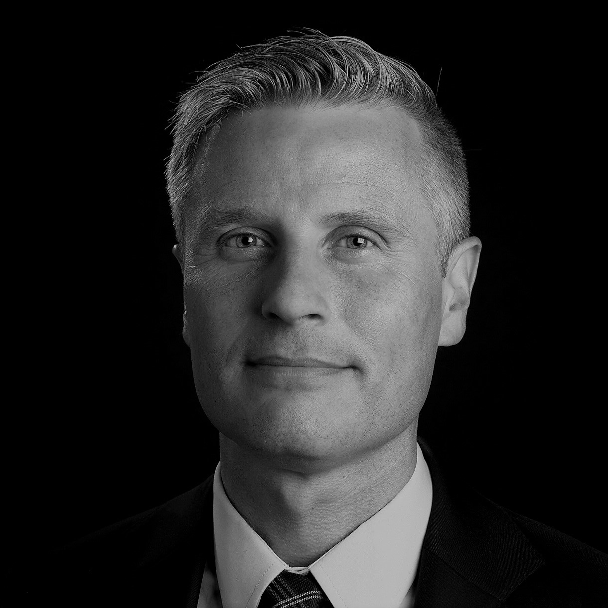 A grayscale image of Claes de Vreese in formal attire, with his face obscured by a gray rectangle. He is wearing a suit jacket, white shirt, and tie, set against a completely black background.