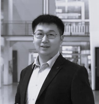 Di Wu, iført briller og jakkesæt, står indendørs. Baggrunden viser moderne arkitektur og slørede strukturelle elementer.