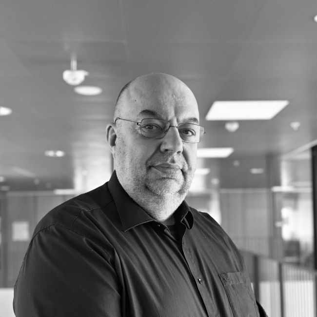 Professor Morten Birkved is standing indoors in a hallway or office space. The background includes ceiling lights, windows, and walls with some visible details of the interior architecture. The picture is in black and white.