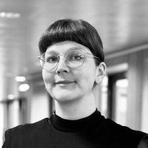 En person med briller og en mørk overdel står indendørs. Baggrunden viser lys og vinduer.