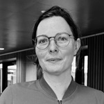 Sort-hvidt billede af en person med håret i hestehale, iført en jakke med lynlås. Baggrunden viser en indendørs gang med lys og vægge.