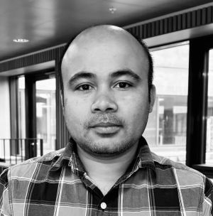 En person iført en ternet skjorte står indendørs. Baggrunden inkluderer store vinduer og en del af loftet med lys.