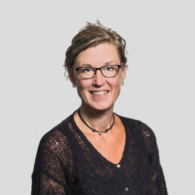 Smilende kvinde med briller iført mørk overdel og halskæde foran grå baggrund.