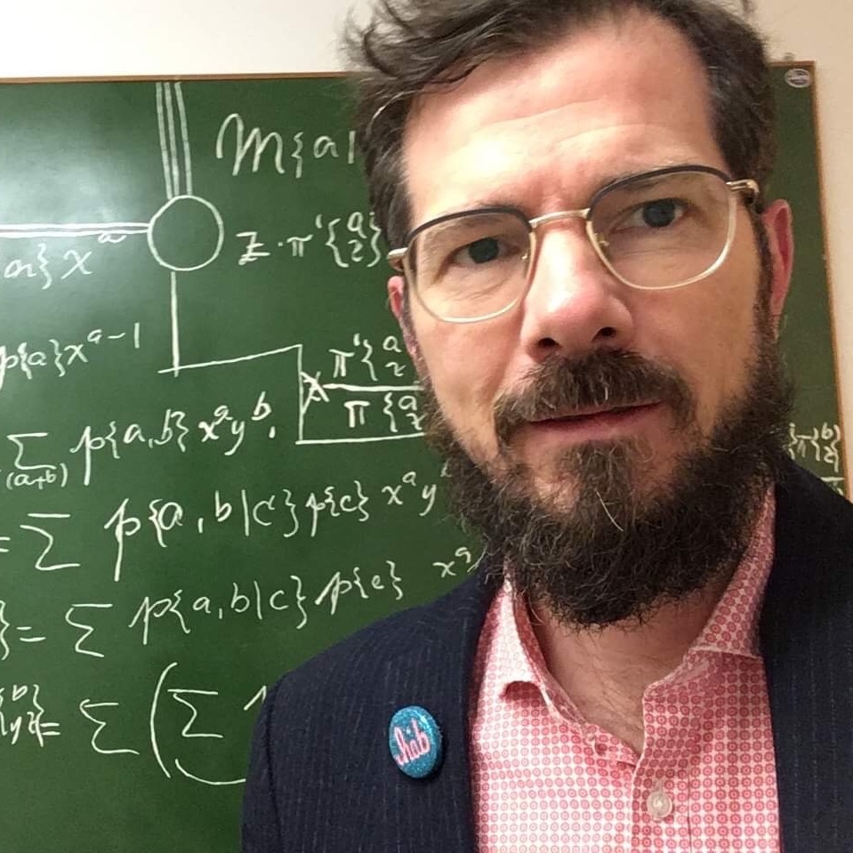 Mand med briller og skæg står foran en grøn tavle med matematiske formler skrevet i hvidt.