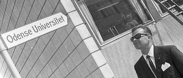 Mand foran bygning med skiltet "Odense Universitet" Sort-hvidt billede af en mand i jakkesæt og solbriller foran en bygning med skiltet "Odense Universitet".