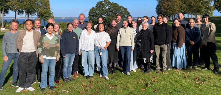 Group photo outdoors at a retreat by the water Large group portrait of retreat attendees standing on grass outdoors near trees and water in sunny weather.