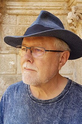 Portrait of an older man wearing a blue hat and glasses Older man with a white beard wearing glasses and a blue hat in front of a stone wall.