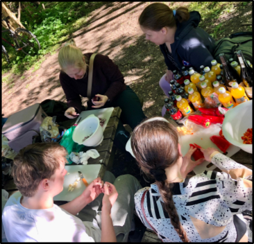 Four people preparing food at a picnic table outdoors with plates, supplies, and assorted bottled drinks.