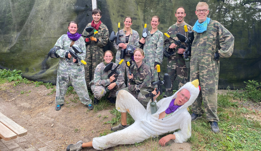 Group of paintball players in camouflage outfits holding markers outdoors, with a person in a white chicken costume lying in front.