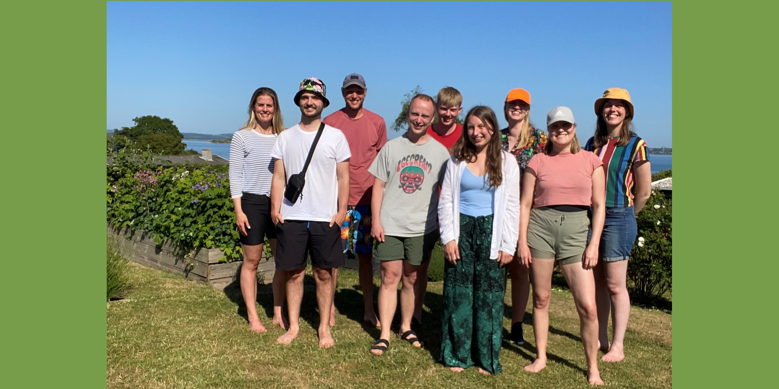 Nine people posing outdoors on a grassy lawn near a garden and waterfront under a clear blue sky in Helnæs, June 2023.