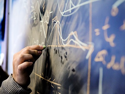 Close-up of a hand drawing diagrams and graph-like markings on a chalkboard.