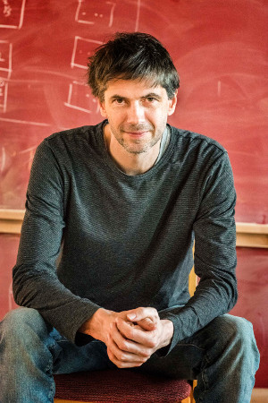 Daniel Merkle seated with clasped hands in front of a red chalkboard.