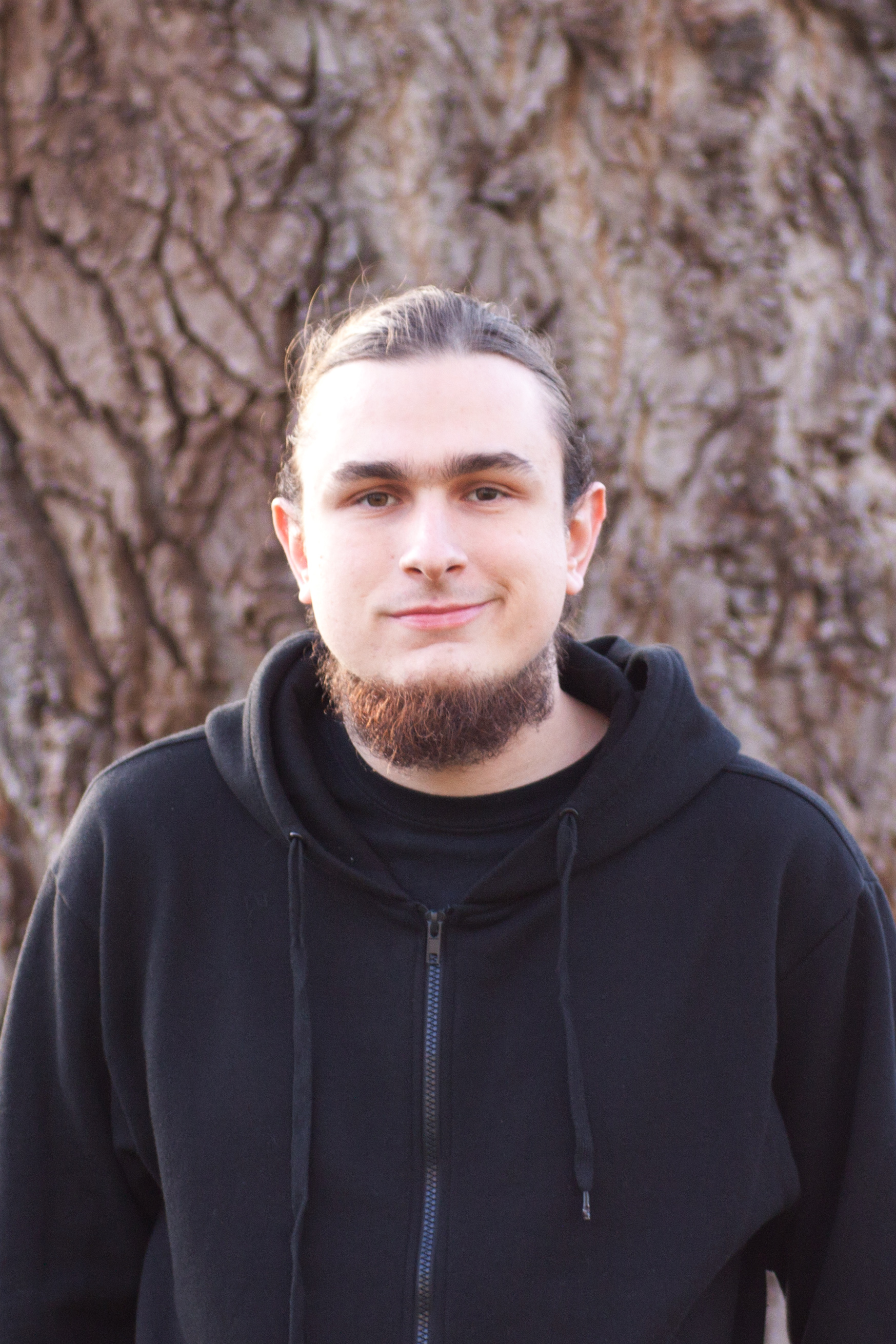 Thomas Gatter wearing a black hoodie and standing in front of a large tree trunk.