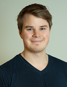 Smiling man in a dark V-neck shirt against a plain light background.