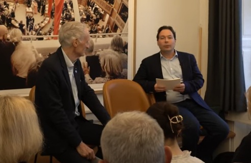 Bertel Haarder and Rasmus Glenthøj participate in a public discussion in a small room with a large poster of the Danish Parliament chamber in the background. The conversation focuses on research from the Nordic Humanities Center.