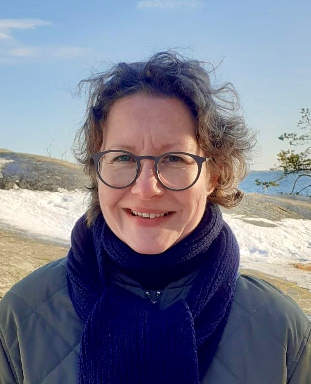 Anu Vehmaa smiling outdoors in a snowy coastal landscape, wearing glasses, a dark scarf, and a jacket.