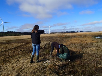 Two people in a grassy field with wind turbines in the background.
