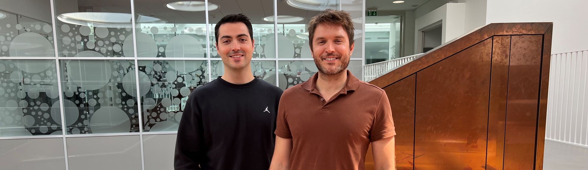 Alvaro and Joel standing together in a modern office atrium with patterned glass walls and a rust-colored staircase.