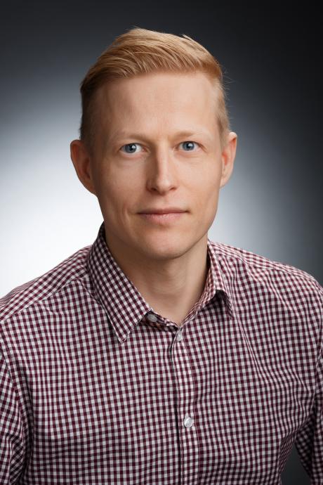 Head-and-shoulders studio portrait of Henrikki Liimatainen in a checkered button-up shirt against a dark gradient background.