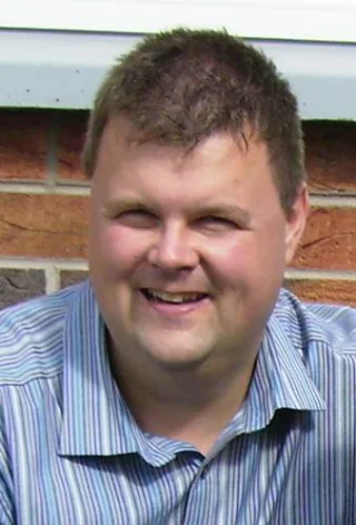 Portrait of John McGrath smiling in a striped collared shirt outdoors.