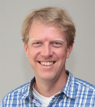 Portrait of Leon Korving smiling in a blue plaid shirt against a gray background.