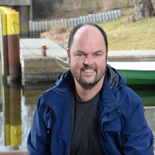 Michael Hupfer smiling outdoors near a waterfront in a blue jacket.