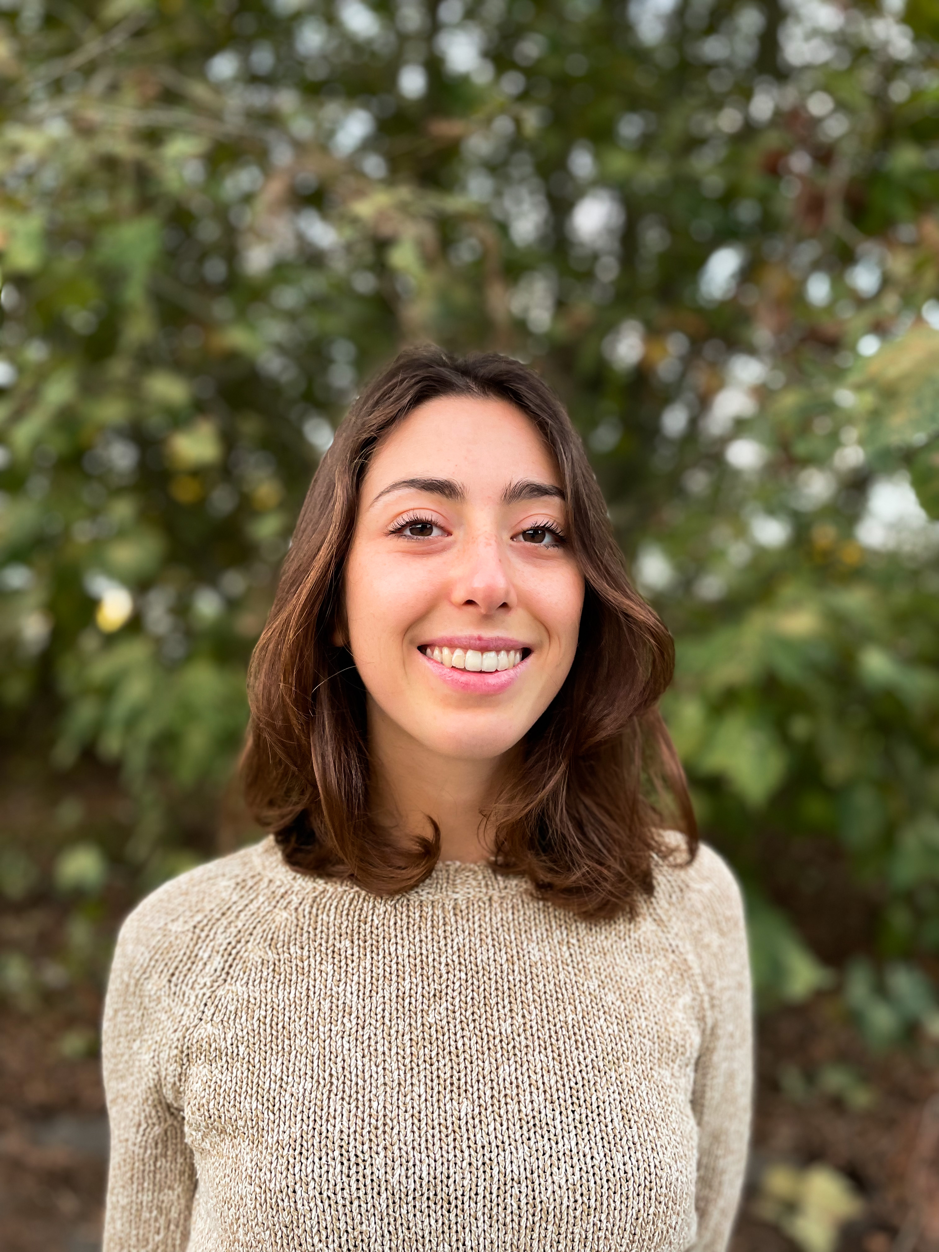 Michela Pacchione smiling in a beige knit sweater outdoors with blurred green foliage behind her.