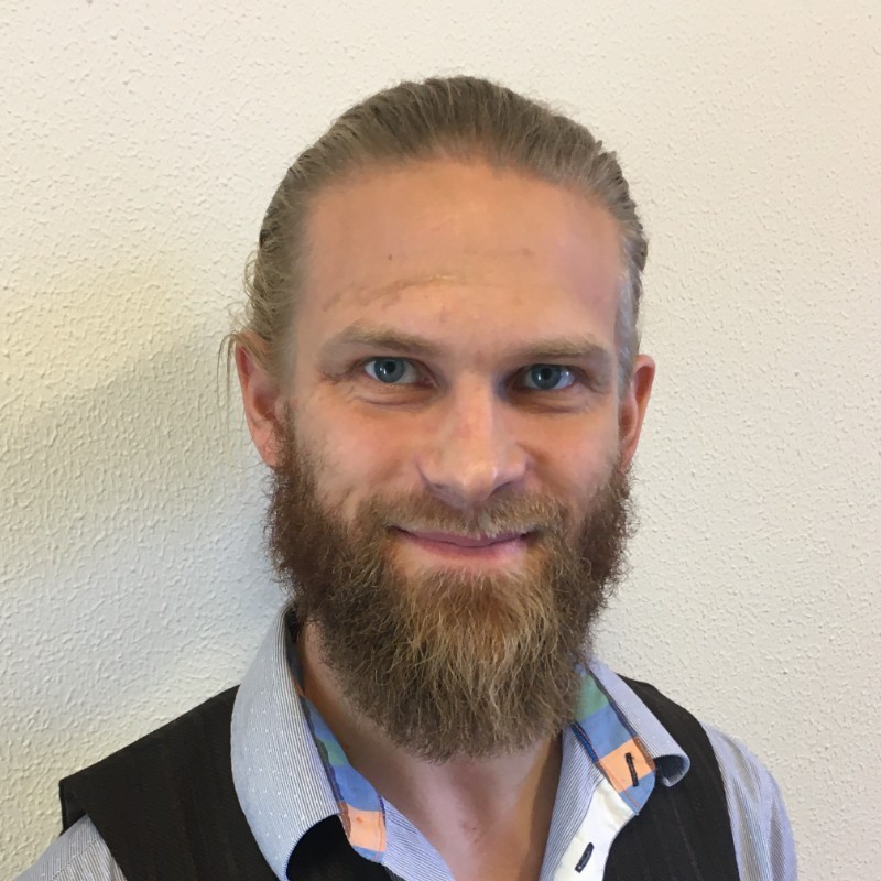 Head-and-shoulders portrait of Mikkel Algren Stokholm-Bjerregaard with tied-back hair and a full beard, smiling in front of a light textured wall.