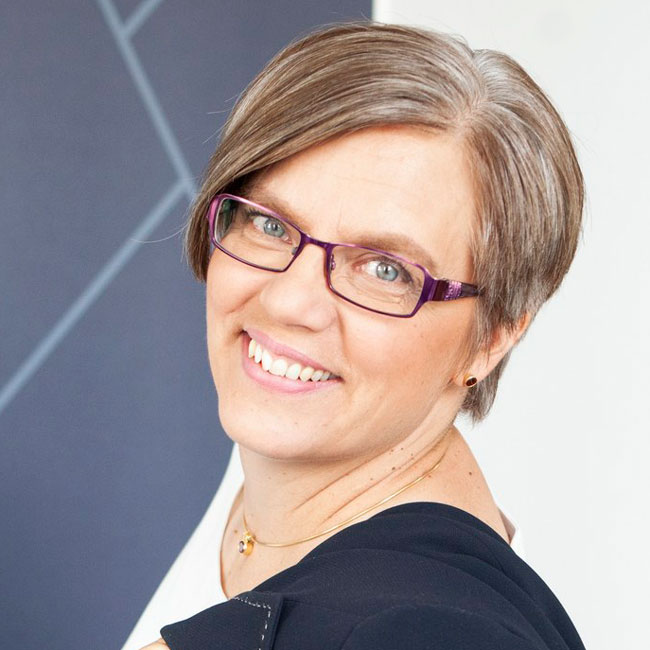 Portrait of Outi Grnfors smiling, wearing purple glasses and a dark top.