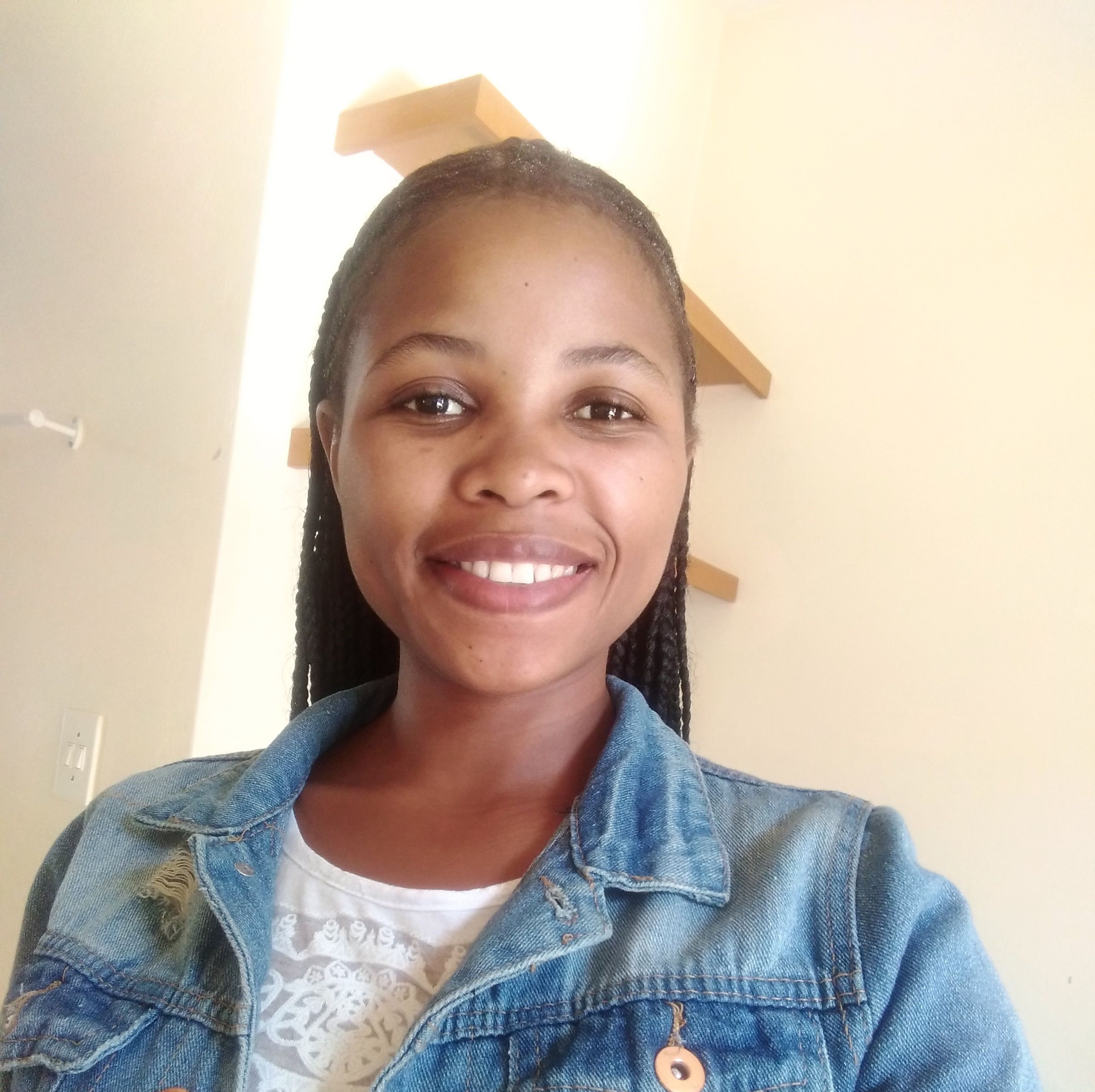 Close-up of a smiling woman with braided hair wearing a blue denim jacket indoors.