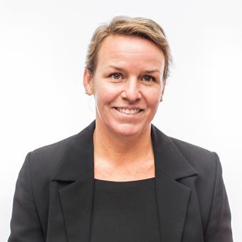 Head-and-shoulders portrait of Tina-Simone Neset in a black blazer and black top against a light gray background.